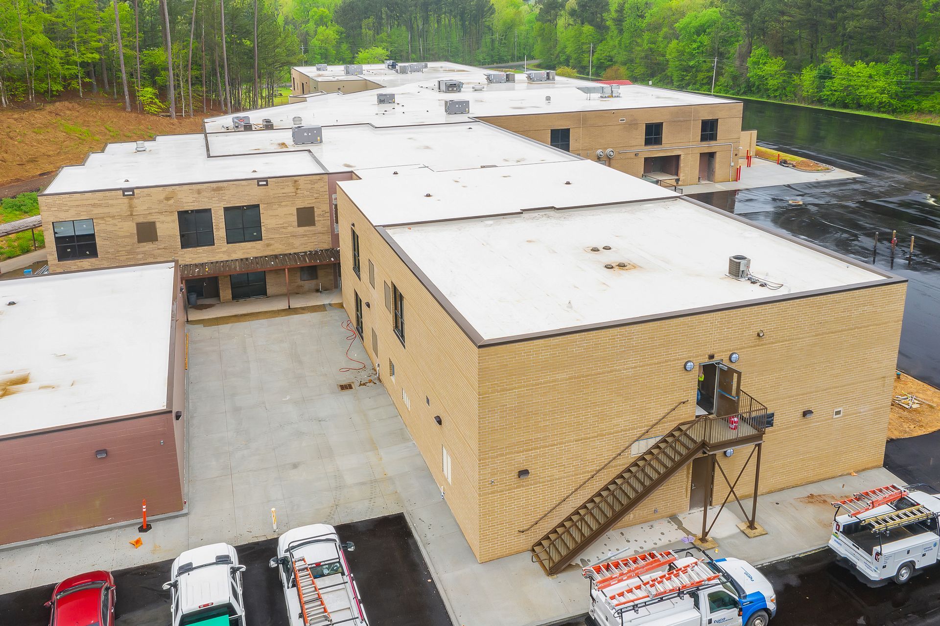 Aerial view of a large beige brick building with a flat white roof and parking lot beside a wooded area