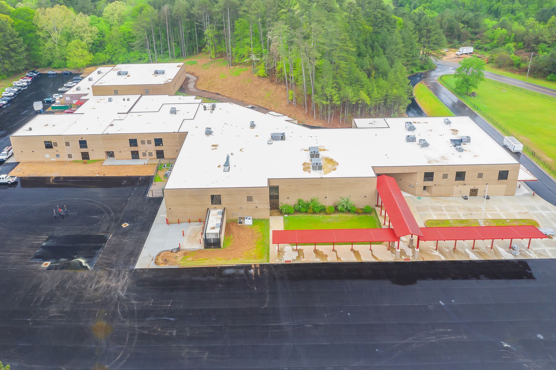 Aerial view of a beige building with a white roof, red awnings, and empty black parking lot surrounded by trees
