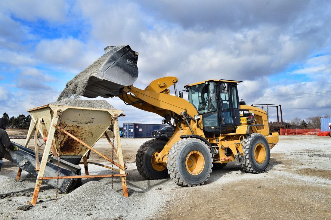 Yellow loader dumping material into a hopper at a construction site. Blue sky overhead.