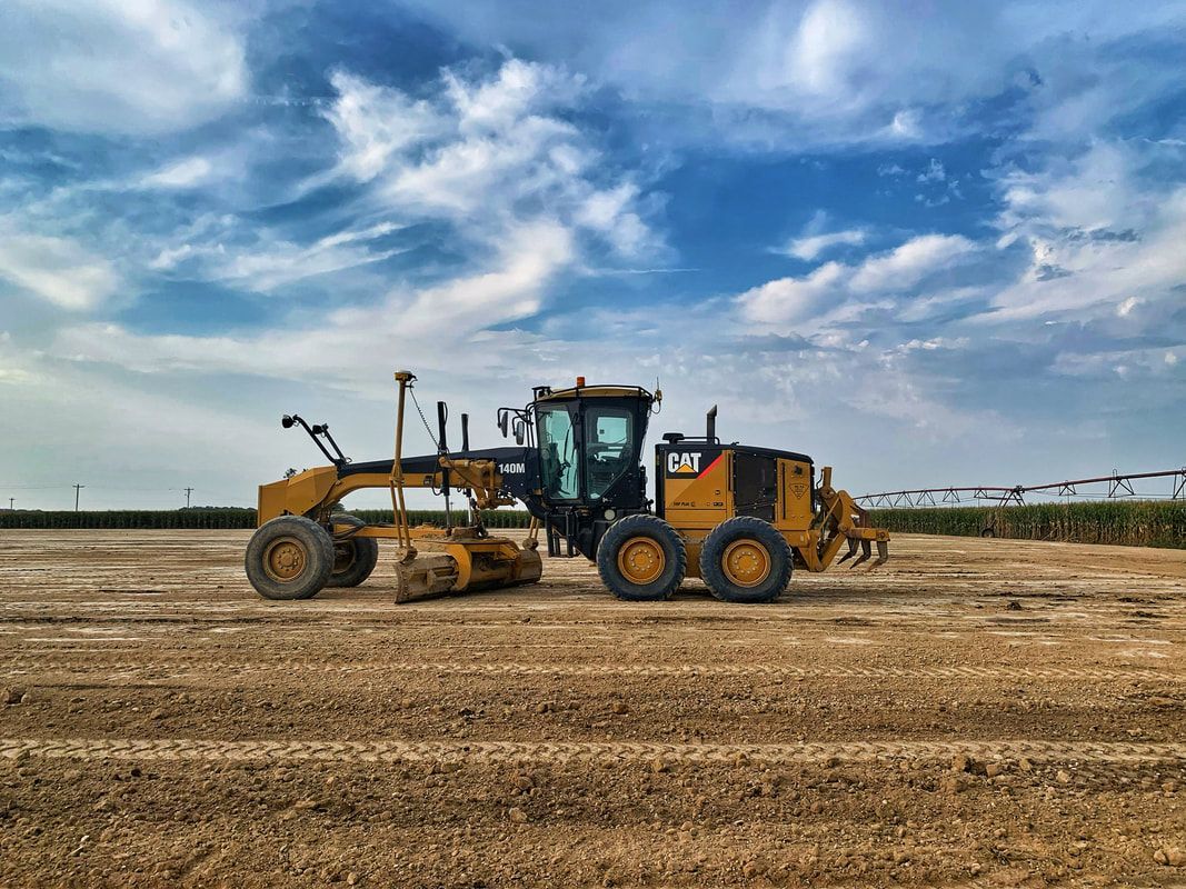 Yellow road grader leveling dirt field under a cloudy blue sky.