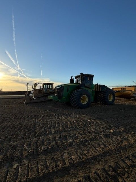 John Deere tractor and bulldozer on a dirt field under a blue sky, with contrails.