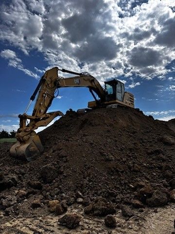 Yellow excavator on a mound of dirt against a blue sky with white clouds.