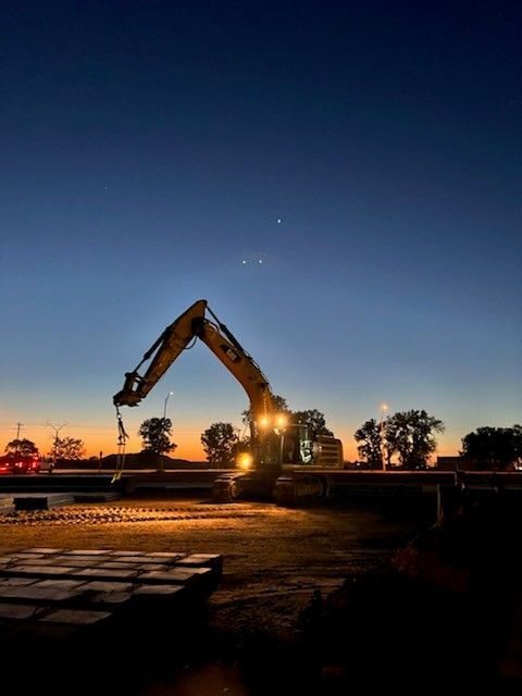 Excavator at dusk, lit up; working on a construction site. Dark blue sky with some stars.