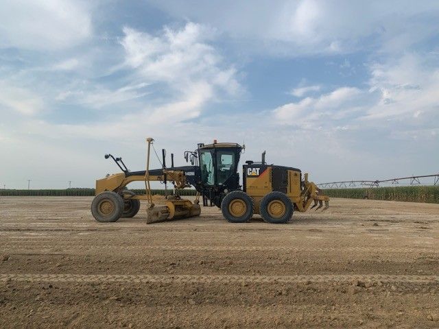 Yellow Caterpillar grader levels dirt in a field under a cloudy sky.