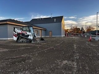 Bobcat tractor on gravel lot in front of a gray building. Construction cones and blue sky visible.