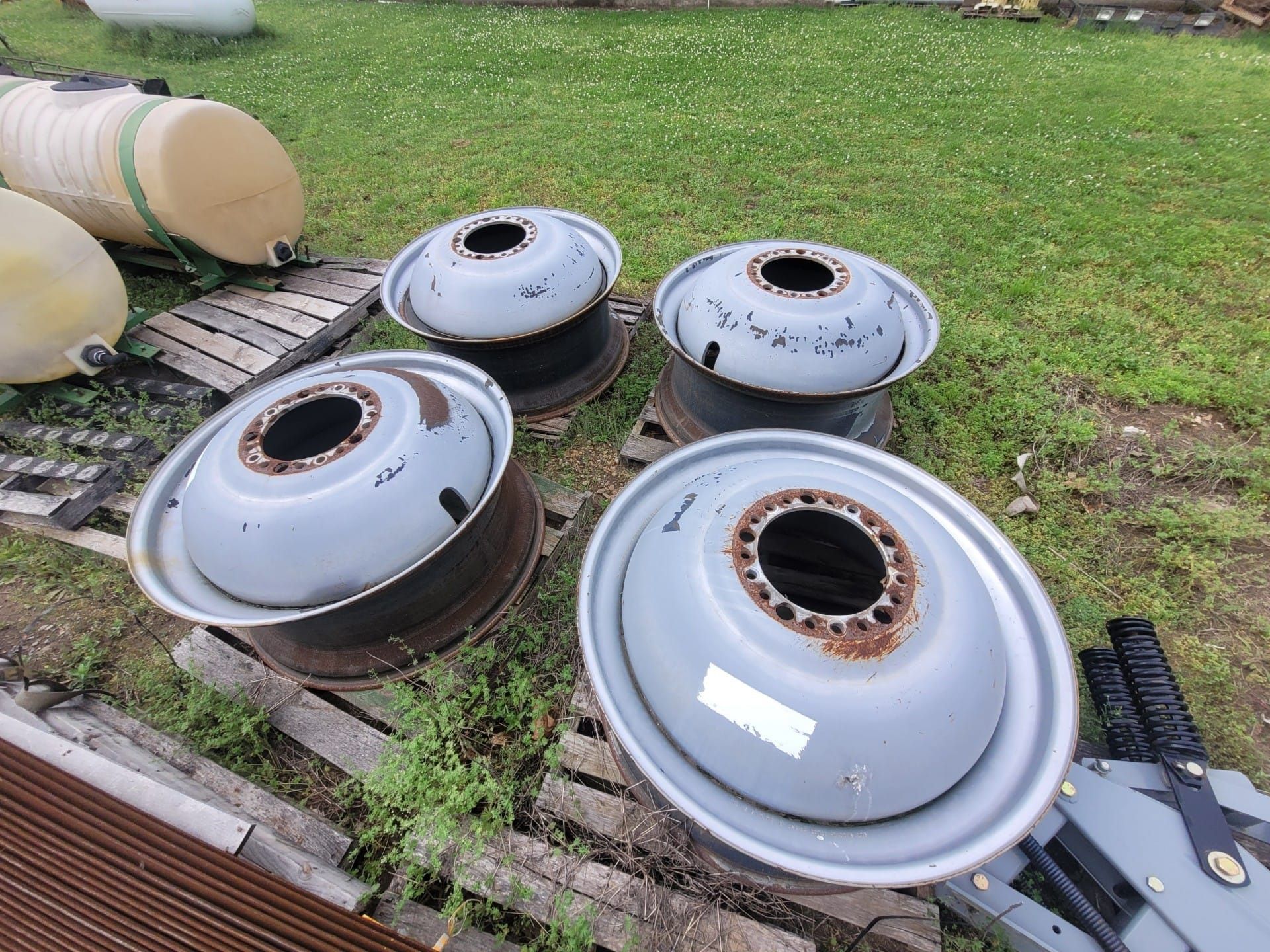 Four gray truck wheels sit on grass, near pallets and tanks.