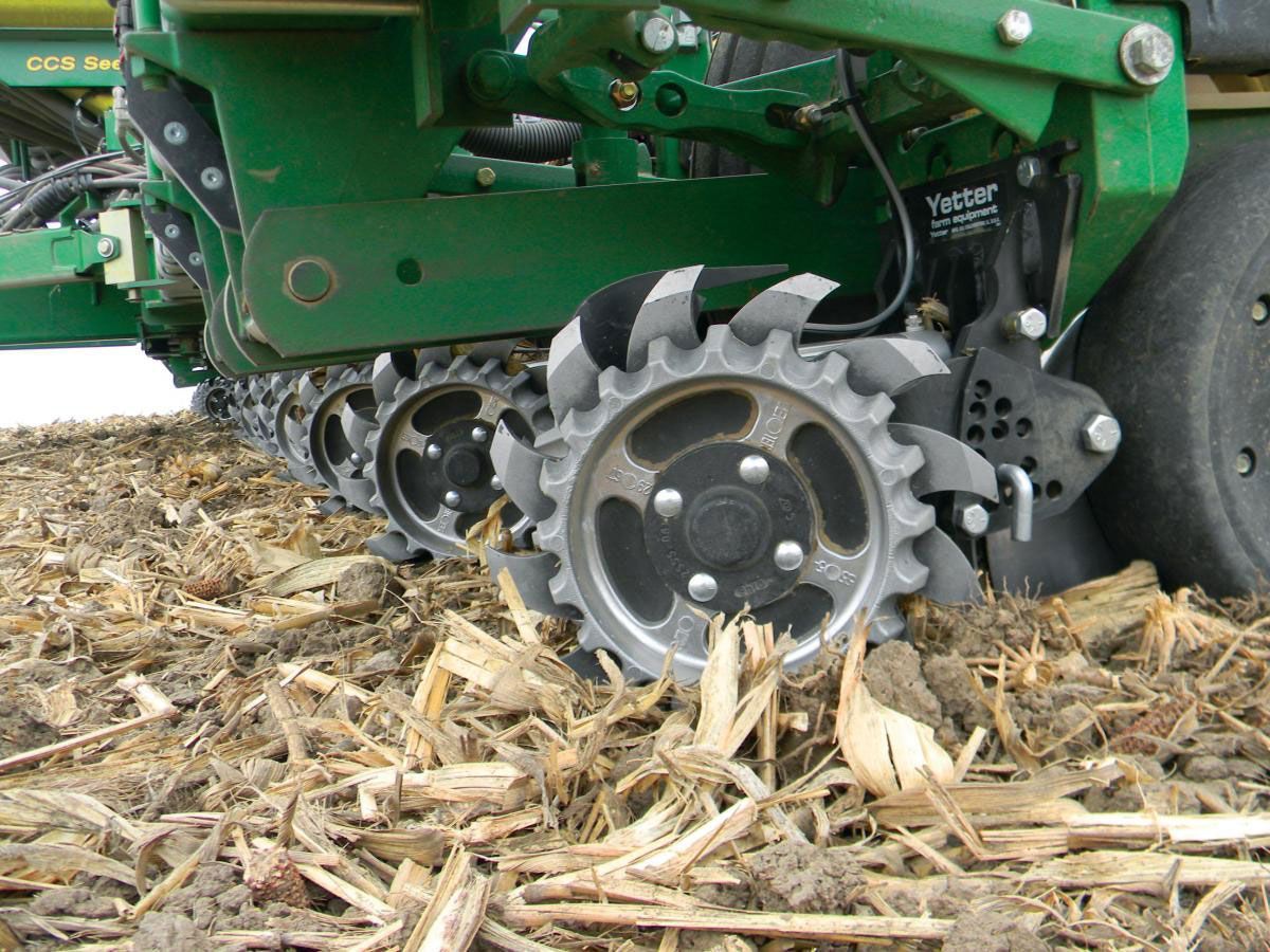 Close-up of a green John Deere planter's row unit with spiked closing wheels in a field of crop residue.