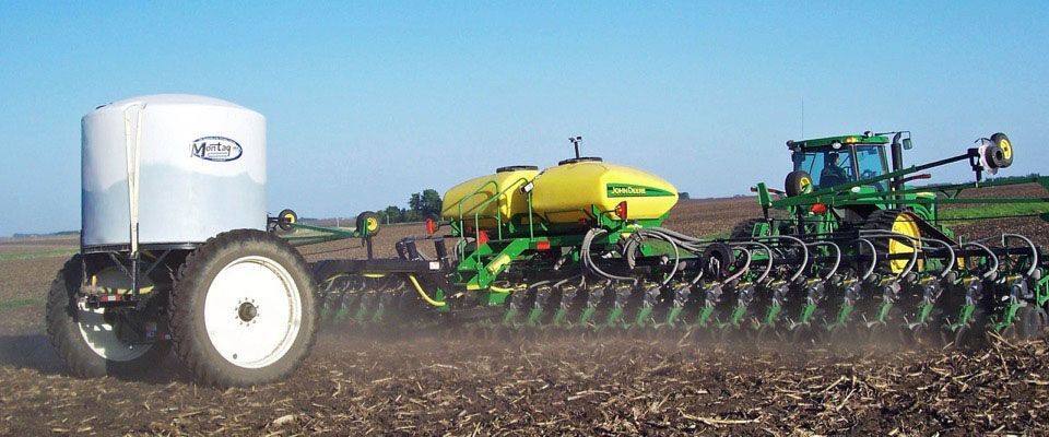 A green tractor pulling a planter across a field, with a tank in tow under a blue sky.