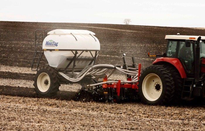A red tractor pulling a white fertilizer applicator across a field, spraying fertilizer.
