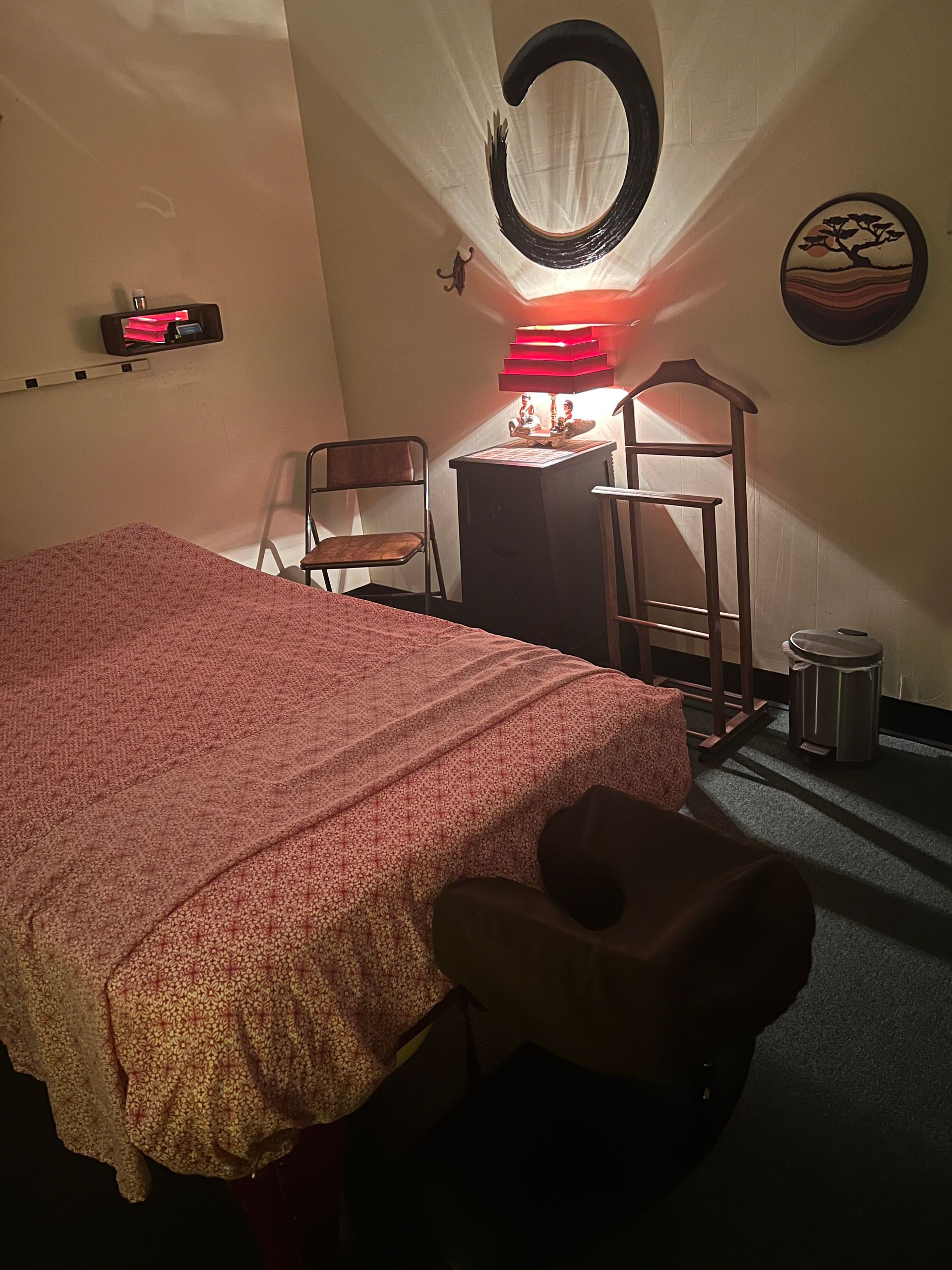 A massage room featuring a treatment table with a headrest, a folding chair, a side table with a lamp, and a valet stand.