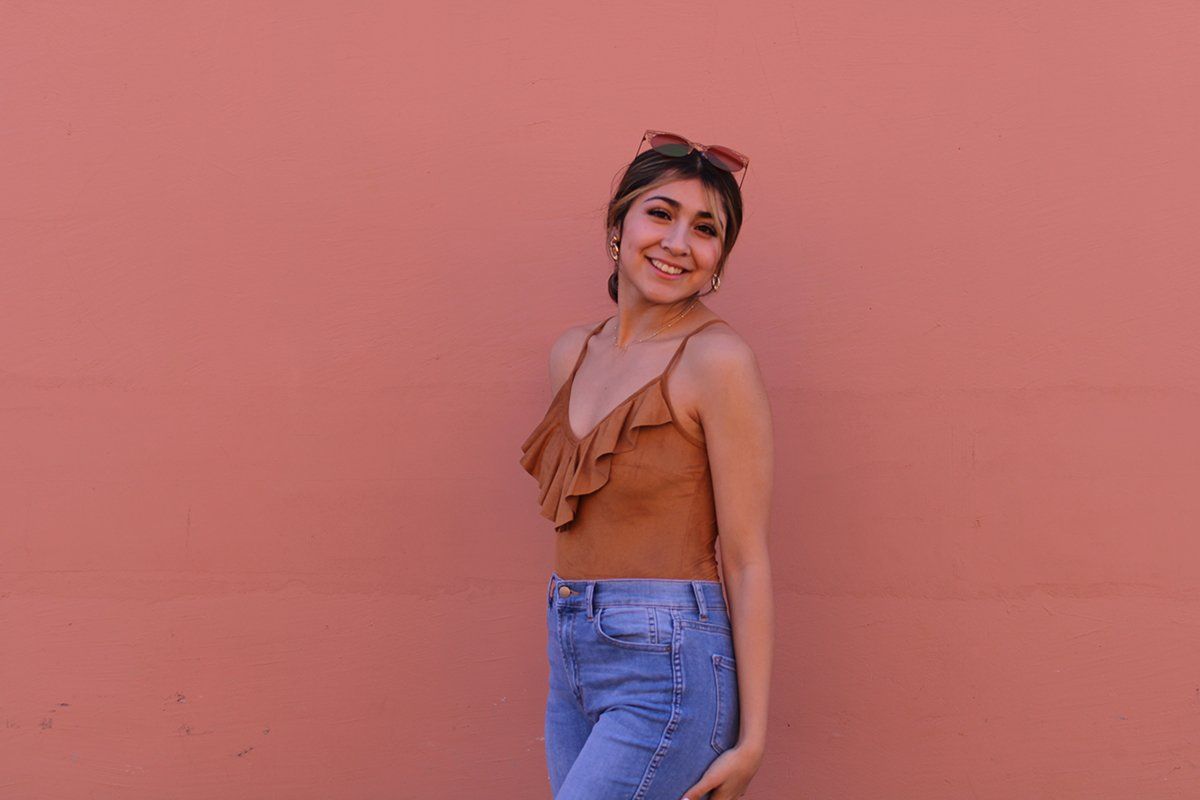 A woman in a brown tank top and blue jeans is standing in front of a pink wall.