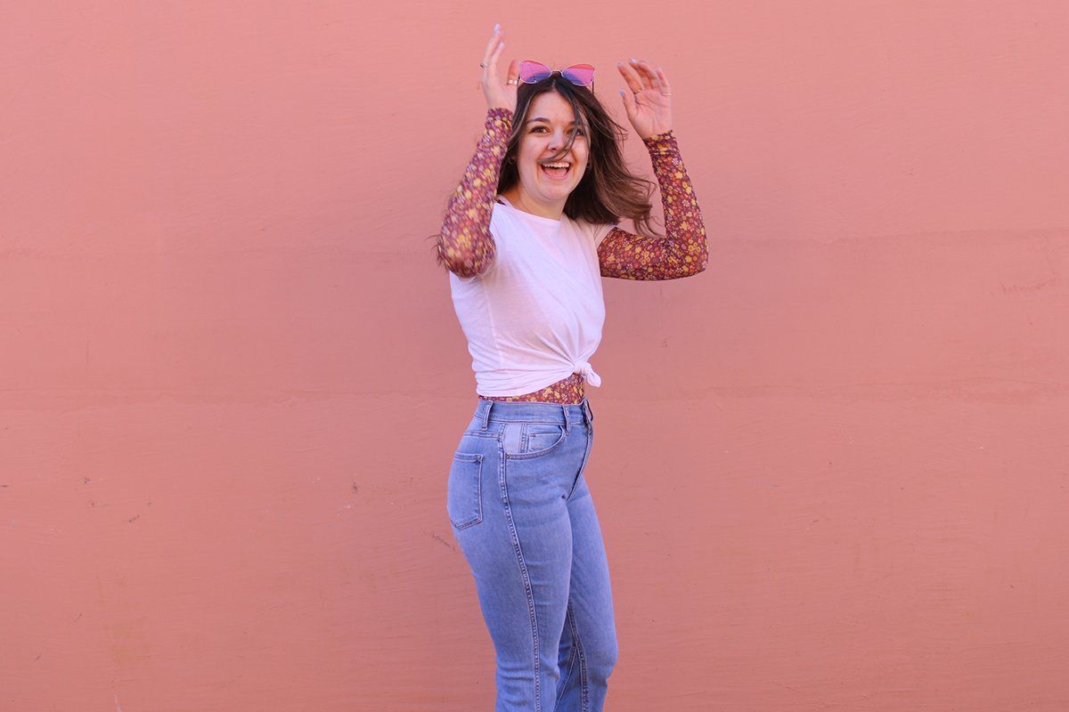 A woman in a white shirt and blue jeans is dancing in front of a pink wall.