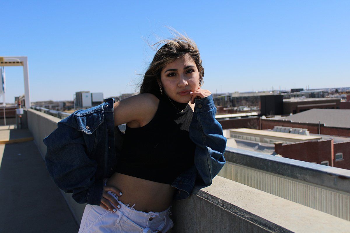 A woman in a crop top and denim jacket is standing on a balcony.