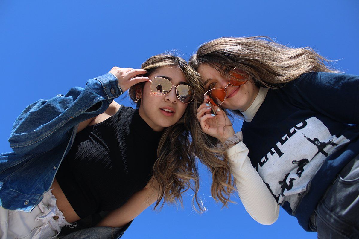 Two girls wearing sunglasses are standing next to each other in front of a blue sky.