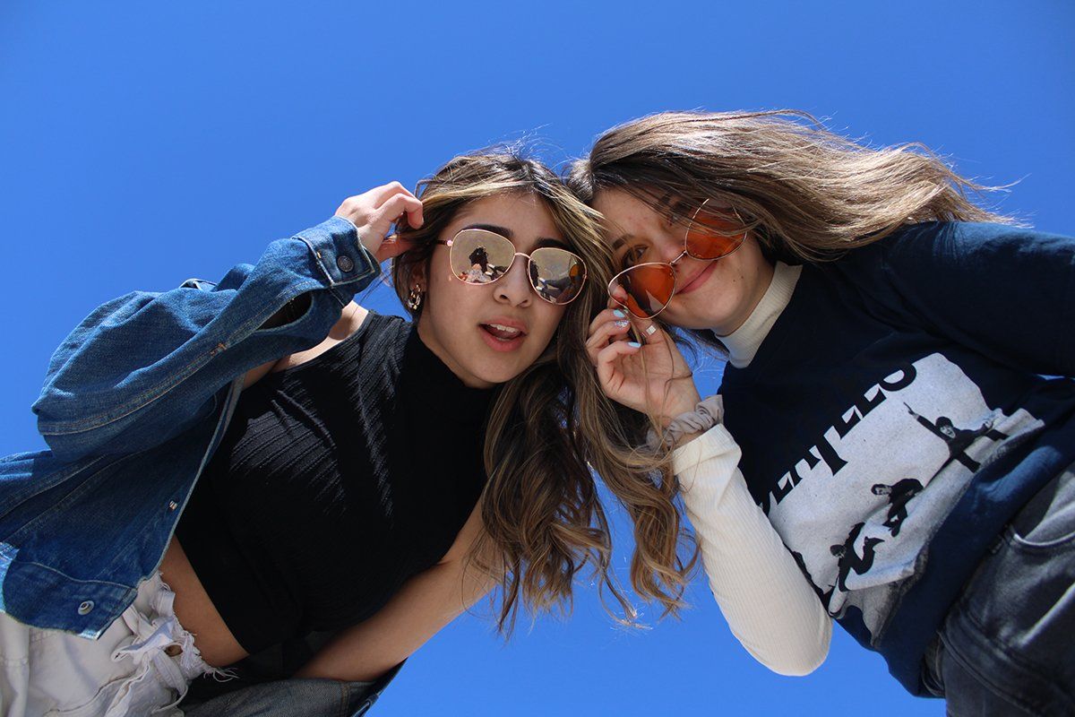 Two girls wearing sunglasses are standing next to each other in front of a blue sky.