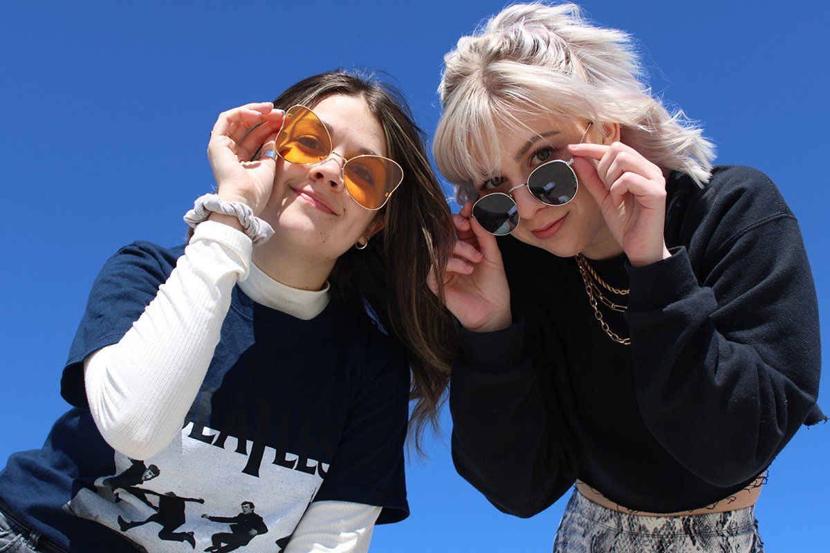 Two girls wearing sunglasses are standing next to each other against a blue sky.