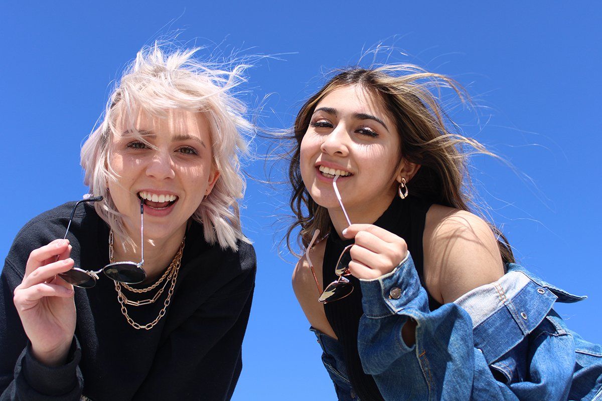 Two women are standing next to each other holding sunglasses in front of a blue sky.