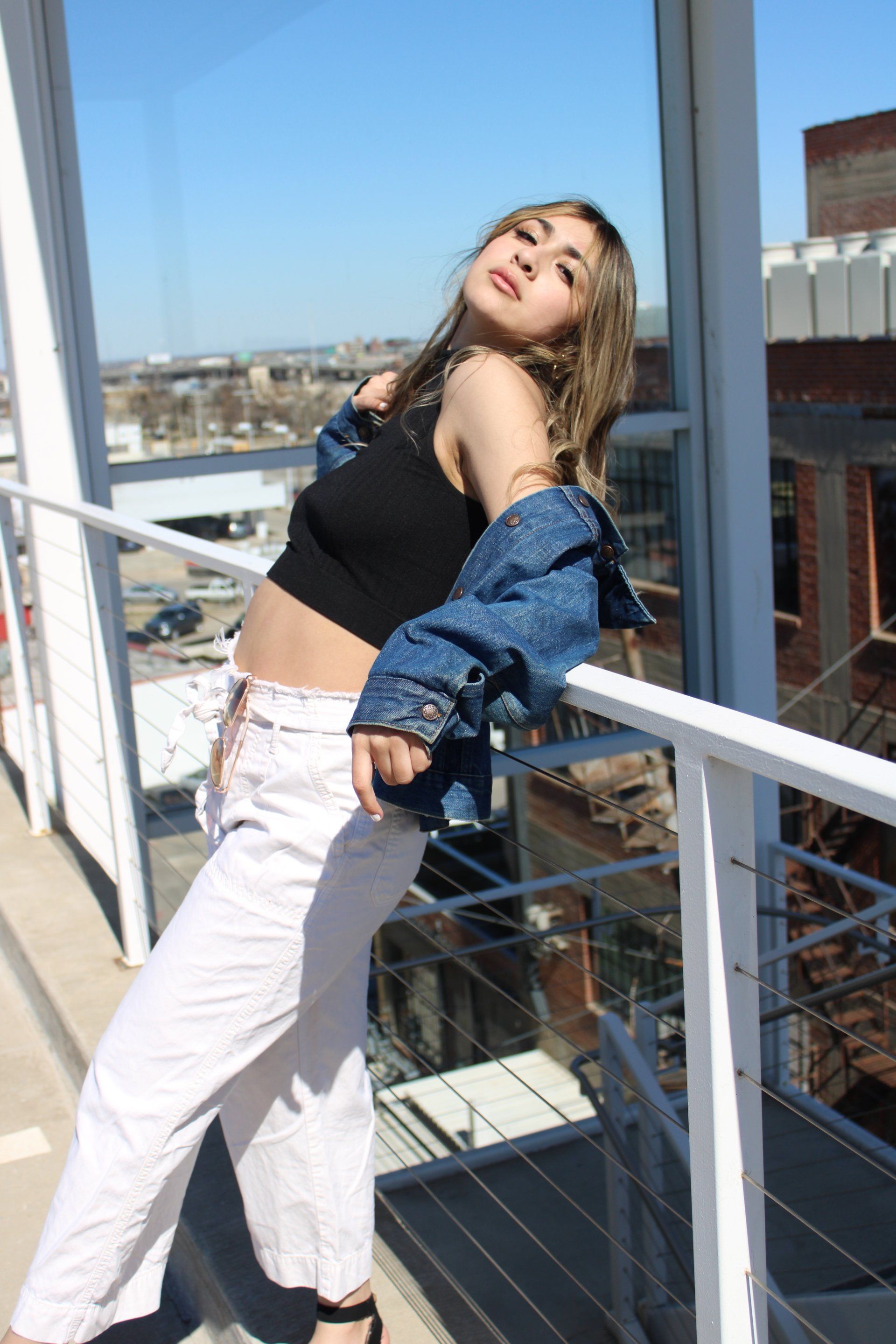 A woman is standing on a balcony wearing a crop top and a denim jacket.