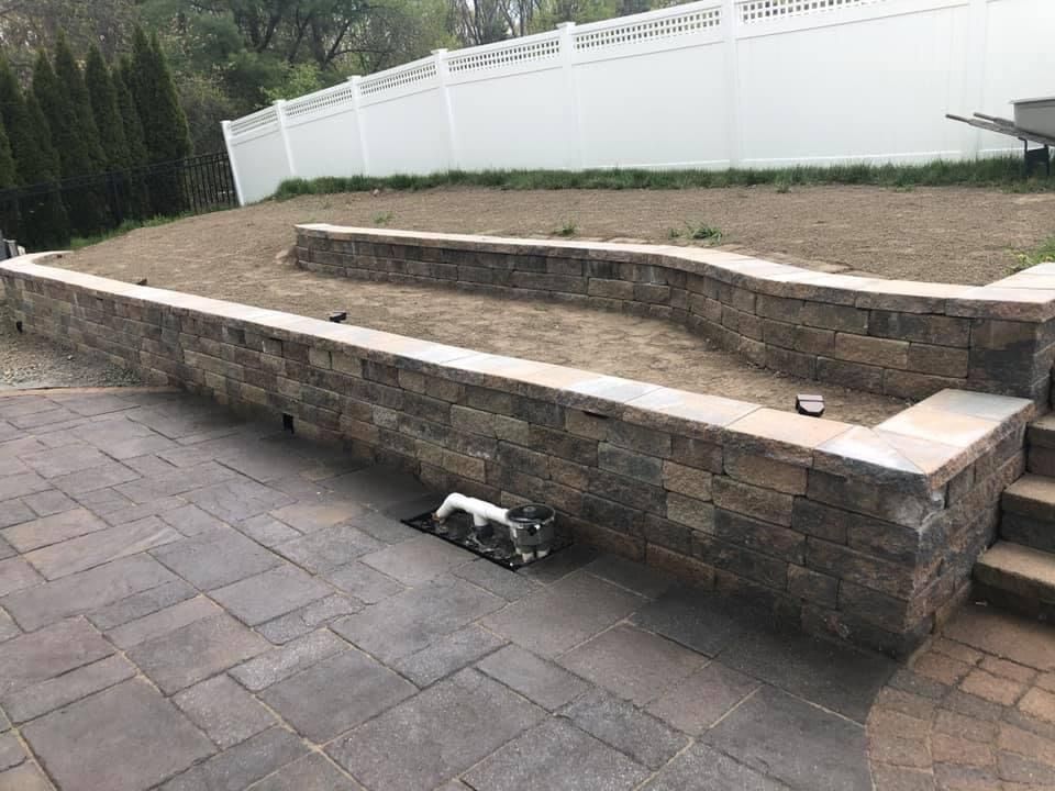 Stone retaining wall with tiered steps beside a paved patio and white fence in a backyard