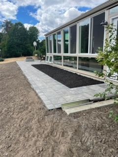 Raised stone patio beside a modern house with large windows, bordered by mulch and a small tree