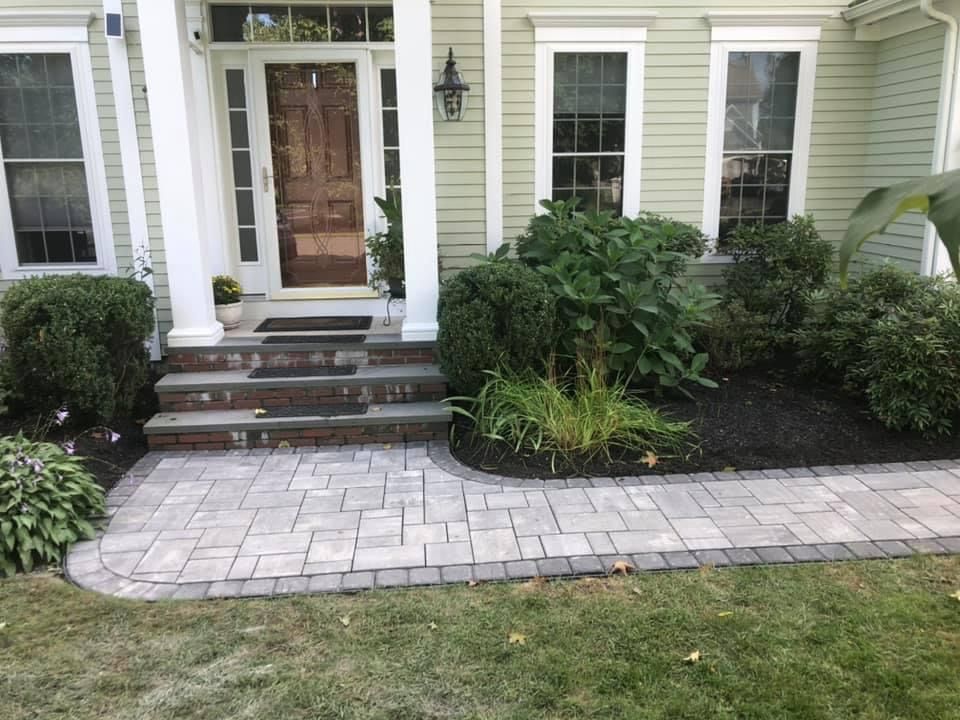 Front entry with stone steps, brick walkway, and shrubs beside a light green house.