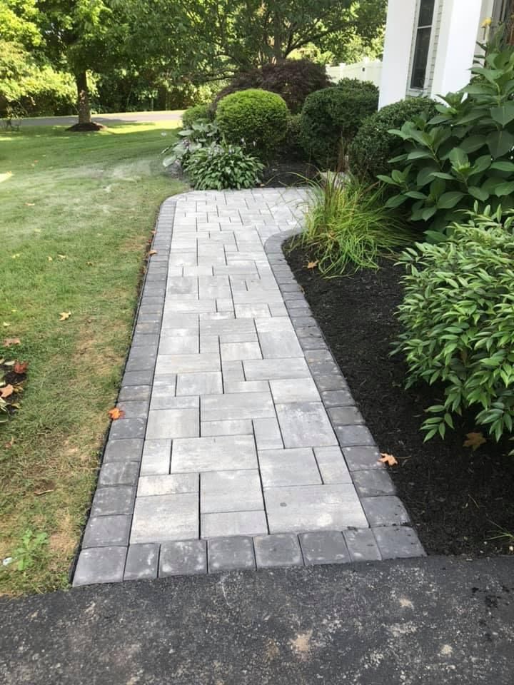 Gray paver walkway beside a lawn and landscaped shrubs leading to a house entrance
