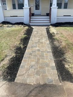 Brick paver walkway leading to a white house’s front steps, bordered by grass.
