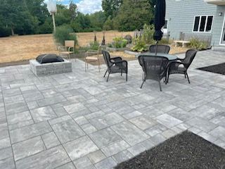 Patio with gray pavers, fire pit, outdoor dining table, chairs, and a closed umbrella beside a house