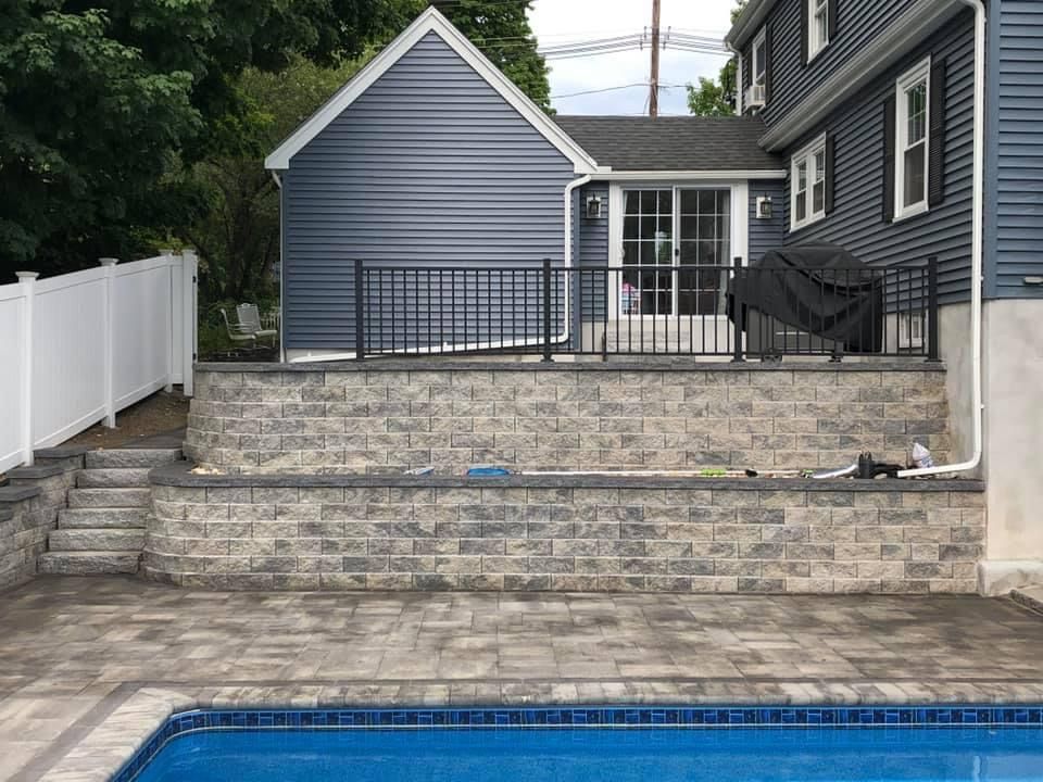 Backyard stone patio with steps, black railing, blue house, and a covered grill beside a pool edge
