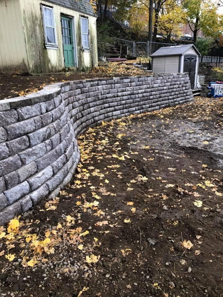 Curved gray stone retaining wall beside a house, with fallen autumn leaves and a small shed in back.