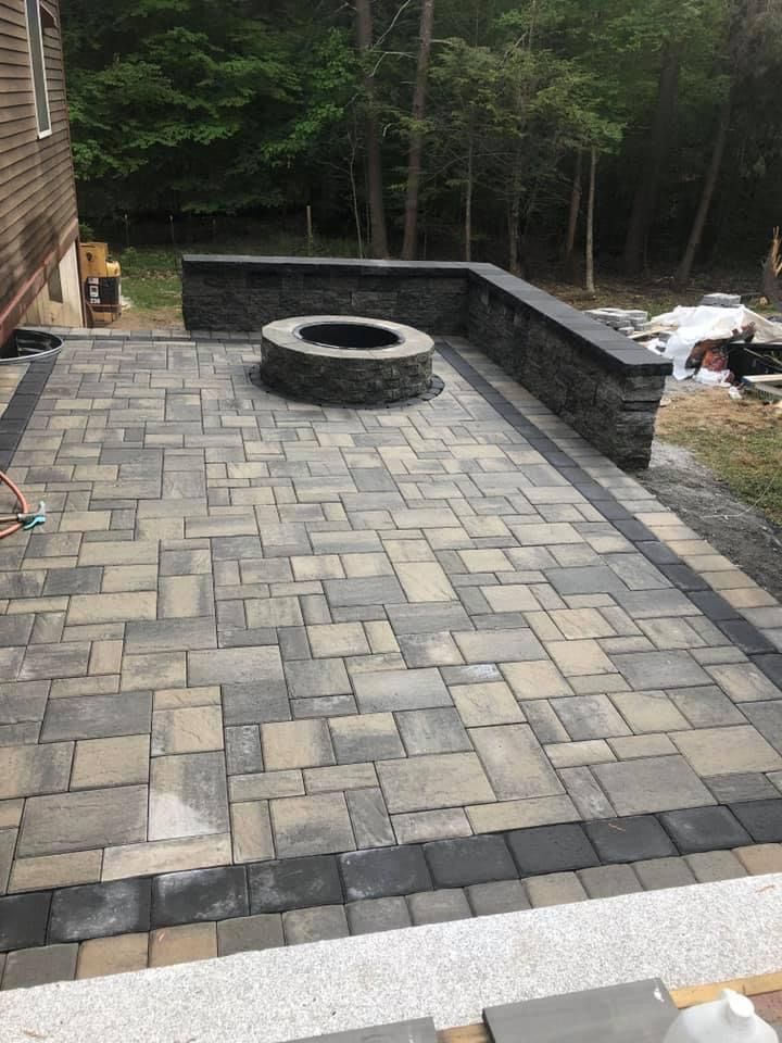 Stone patio with circular fire pit, bordered by a dark retaining wall and woods beyond.
