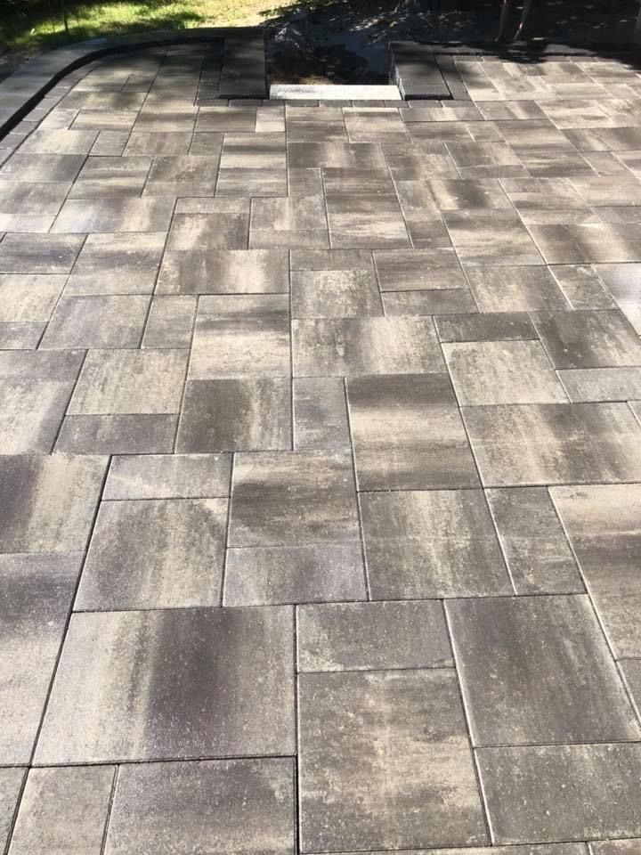 Gray paved patio with rectangular stone tiles in sunlight, bordered by grass and a dark outdoor chair shadow.