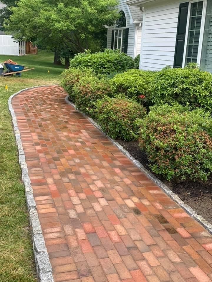 Brick walkway beside a white house, bordered by shrubs and trees in a green yard.