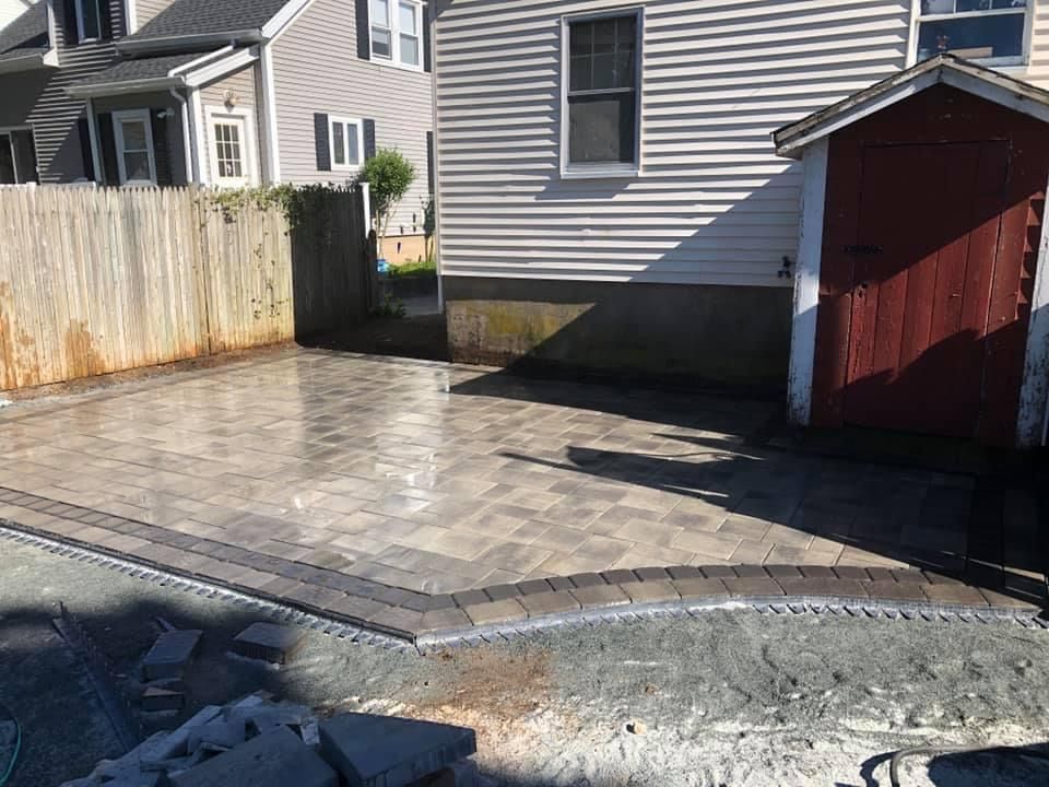 Freshly paved gray patio beside a house, with a red shed and white fence in the background