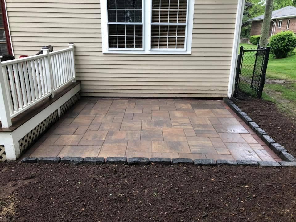 Small brick patio beside a beige house, bordered by a white porch railing and garden bed.