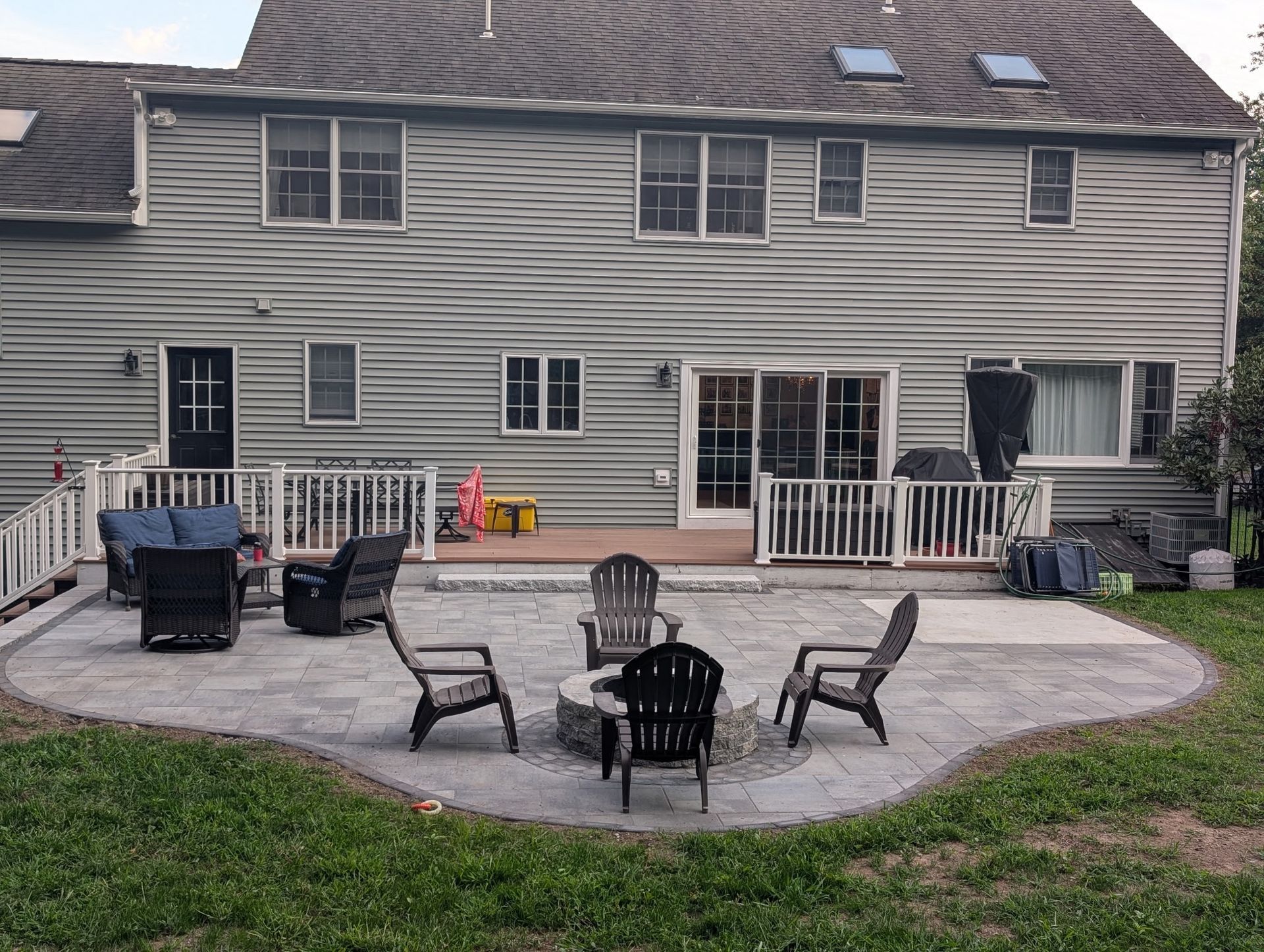 Backyard patio with circular stone fire pit, wicker chairs, grill, and a house deck in the background