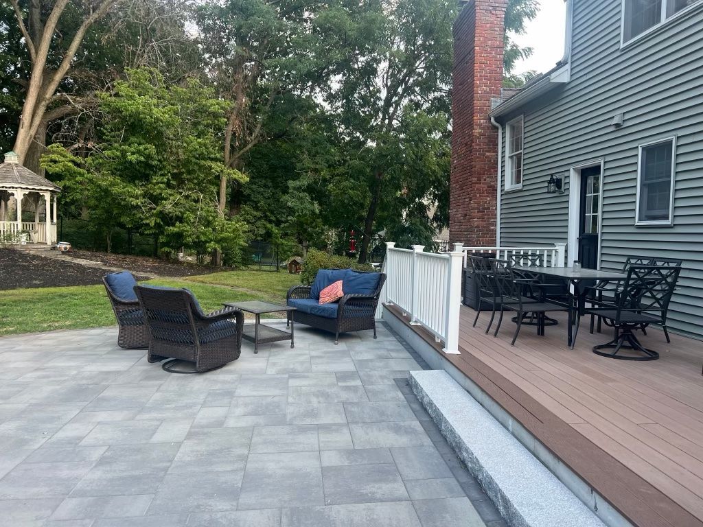 Patio with wicker chairs and table beside a house, overlooking a stone patio and wooded yard
