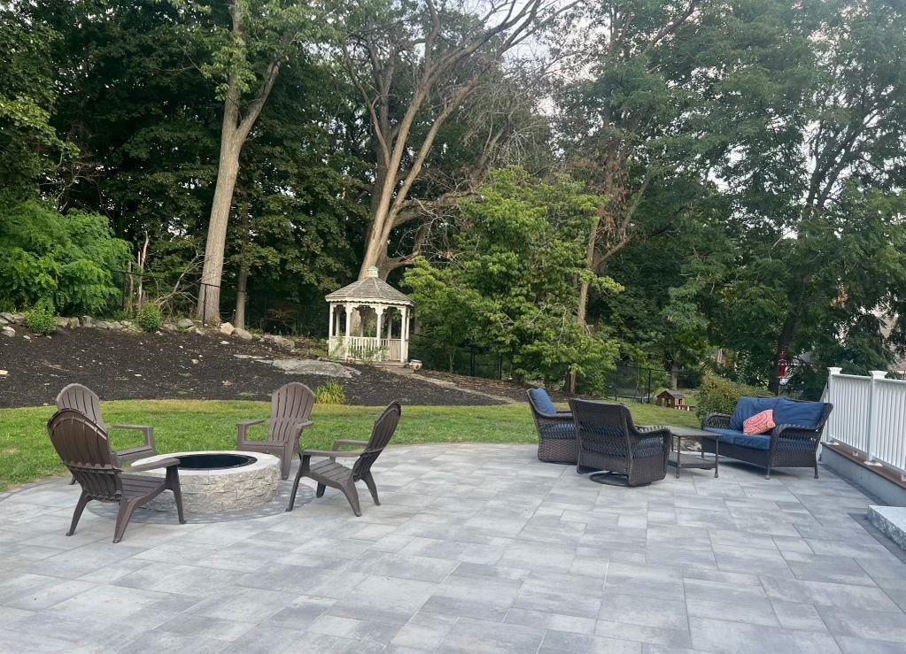 Outdoor patio with wicker chairs, tables, and a gray stone terrace beside trees and greenery