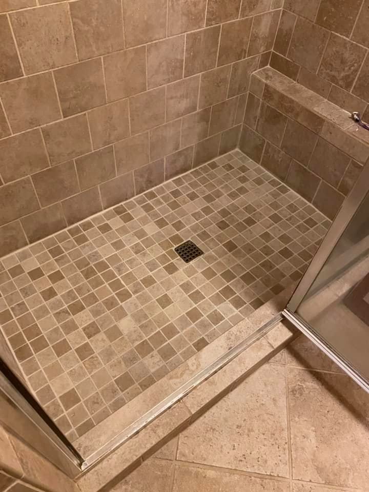 Tiled shower with a low curb and small square tiles. A drain sits in the center. A glass door is visible.