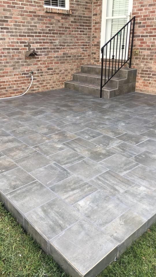 Brick patio with gray pavers and steps leading to a door with a black railing, set against a brick wall.