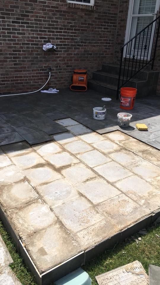 Patio construction: laying gray paving stones on a mortar bed. Tools and materials scattered.