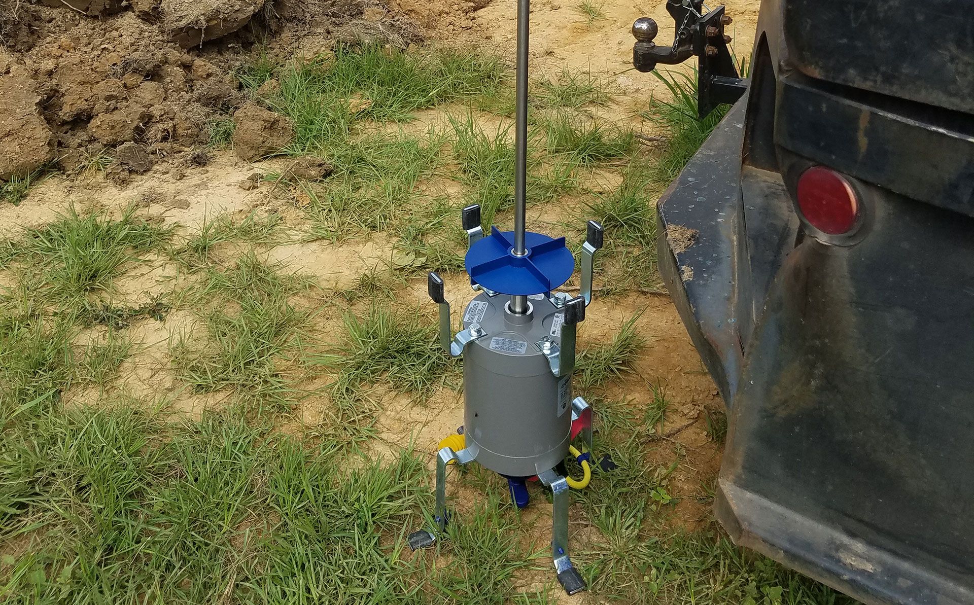 Robot next to a truck. It is grey, blue, and silver, on grass. The robot has legs, a mast, and a blue top.