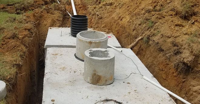 A septic tank with open concrete manholes in a dirt trench, surrounded by white PVC pipes.