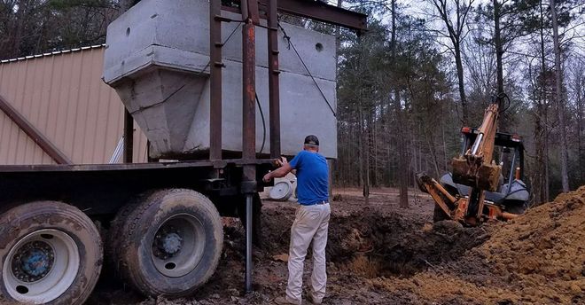 Man working near a truck with large concrete structure being lowered into a hole by backhoe in wooded area.
