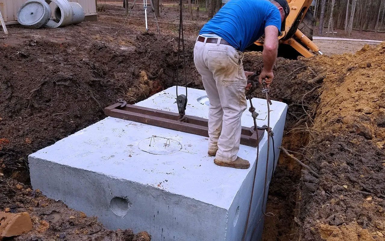 Man lowering a concrete septic tank into an excavated trench.