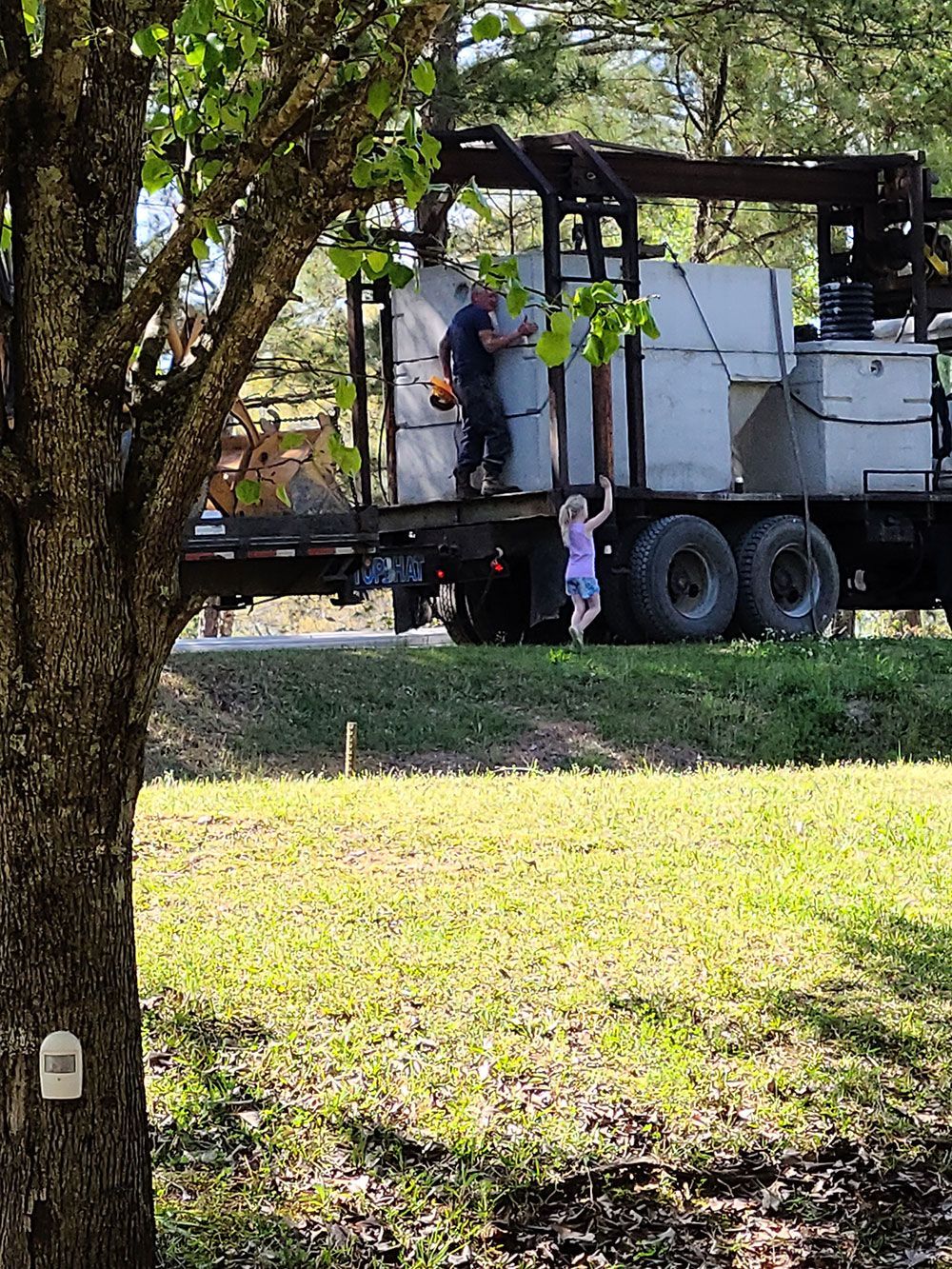 A person and a child on a truck unloading large gray boxes in a grassy area with trees.