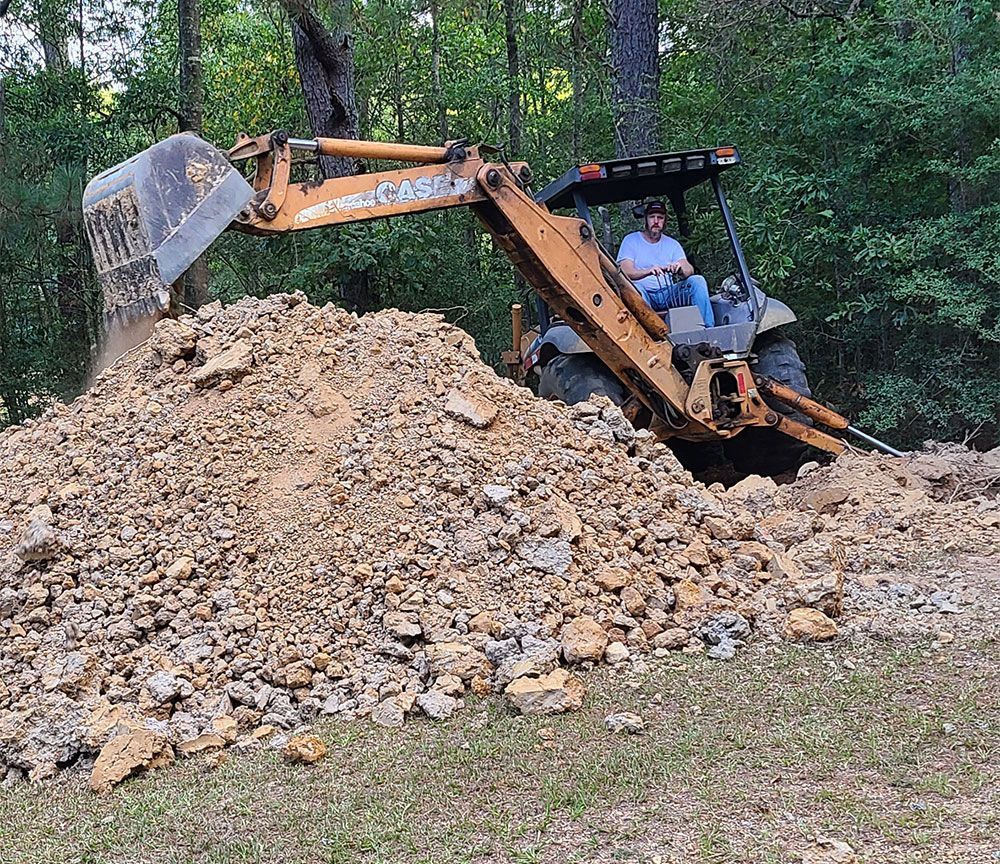 A man operating a Case backhoe loads dirt into a pile outdoors, under trees.