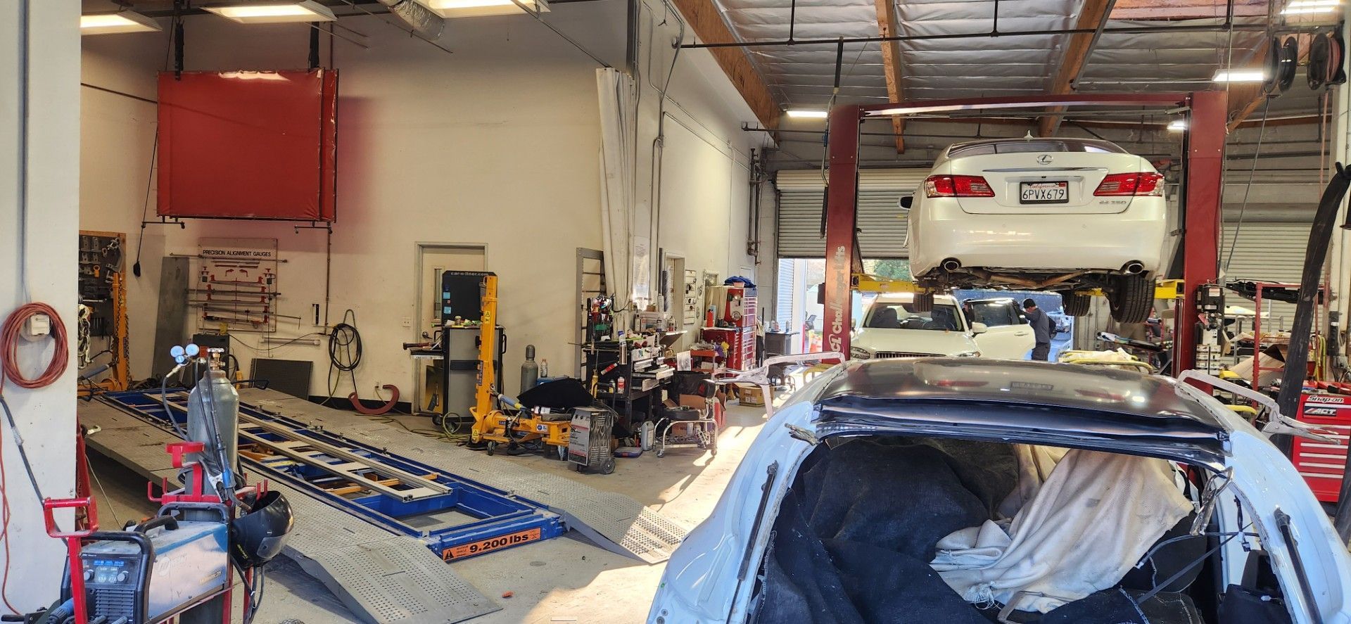 Inside a car repair shop, a car is on a lift, and another damaged vehicle is in the foreground.
