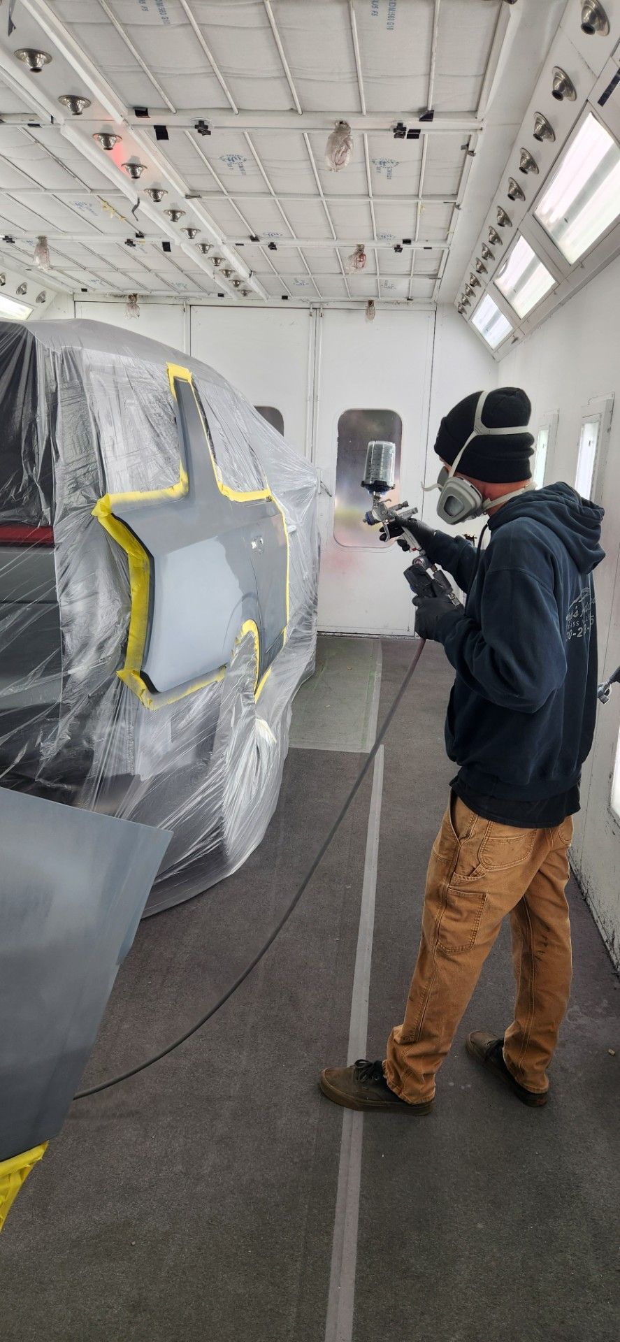 A person in protective gear sprays a vehicle panel in a paint booth.