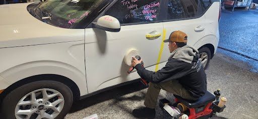 A person polishing the side of a white car, kneeling on a small red cart.